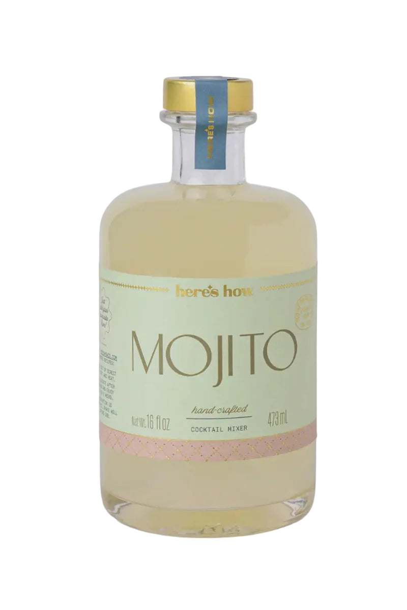 A bottle of Here's How Mojito cocktail mixer, featuring a pale green label with gold lettering and a pink band near the bottom, sealed with a gold cap and blue neck seal, on a light gray background.
